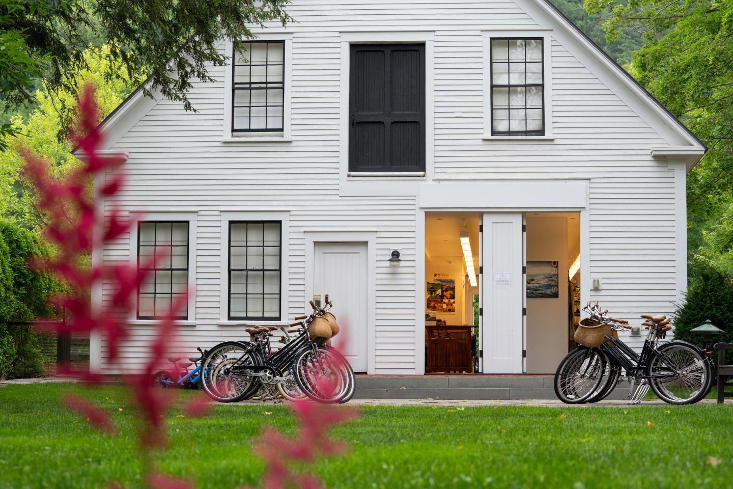 White barn with open door, bicycles in front, surrounded by greenery.
