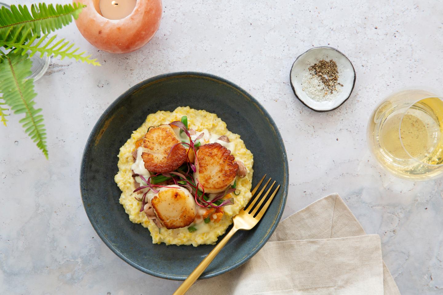 Seared scallops on risotto with a golden fork, candle, and a drink nearby.