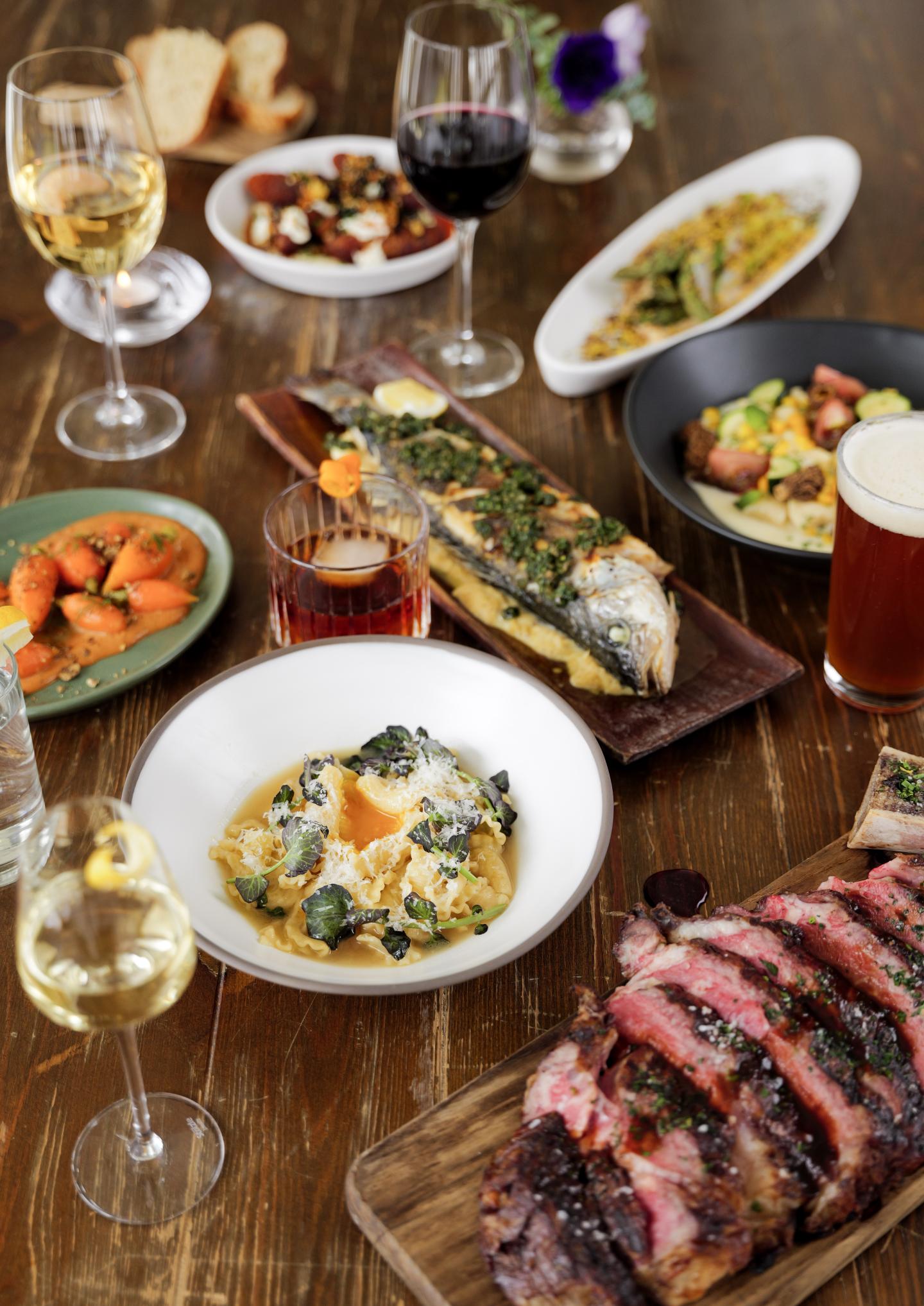 Gourmet meal with steak, pasta, fish, and wine on a wooden table.