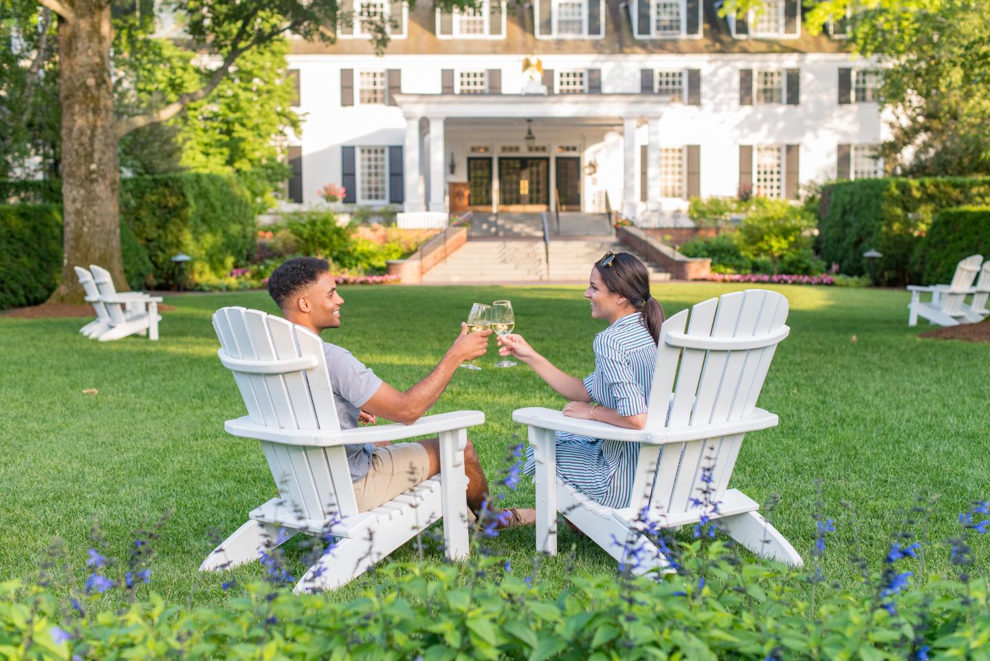A couple clinks glasses on lawn chairs in front of a large white house.