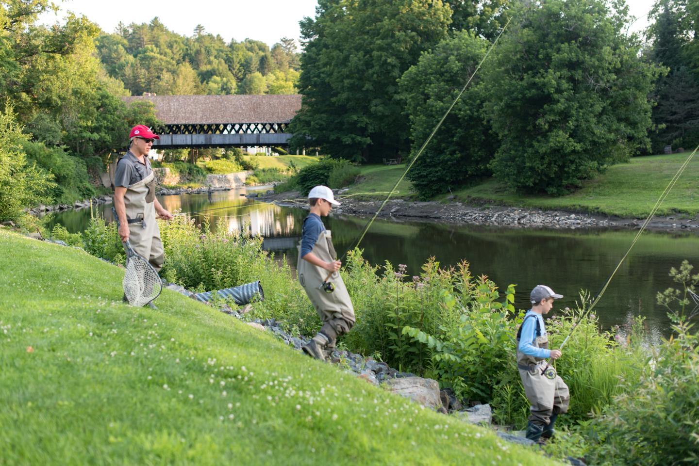Three people fishing by a river, with a covered bridge in the background.