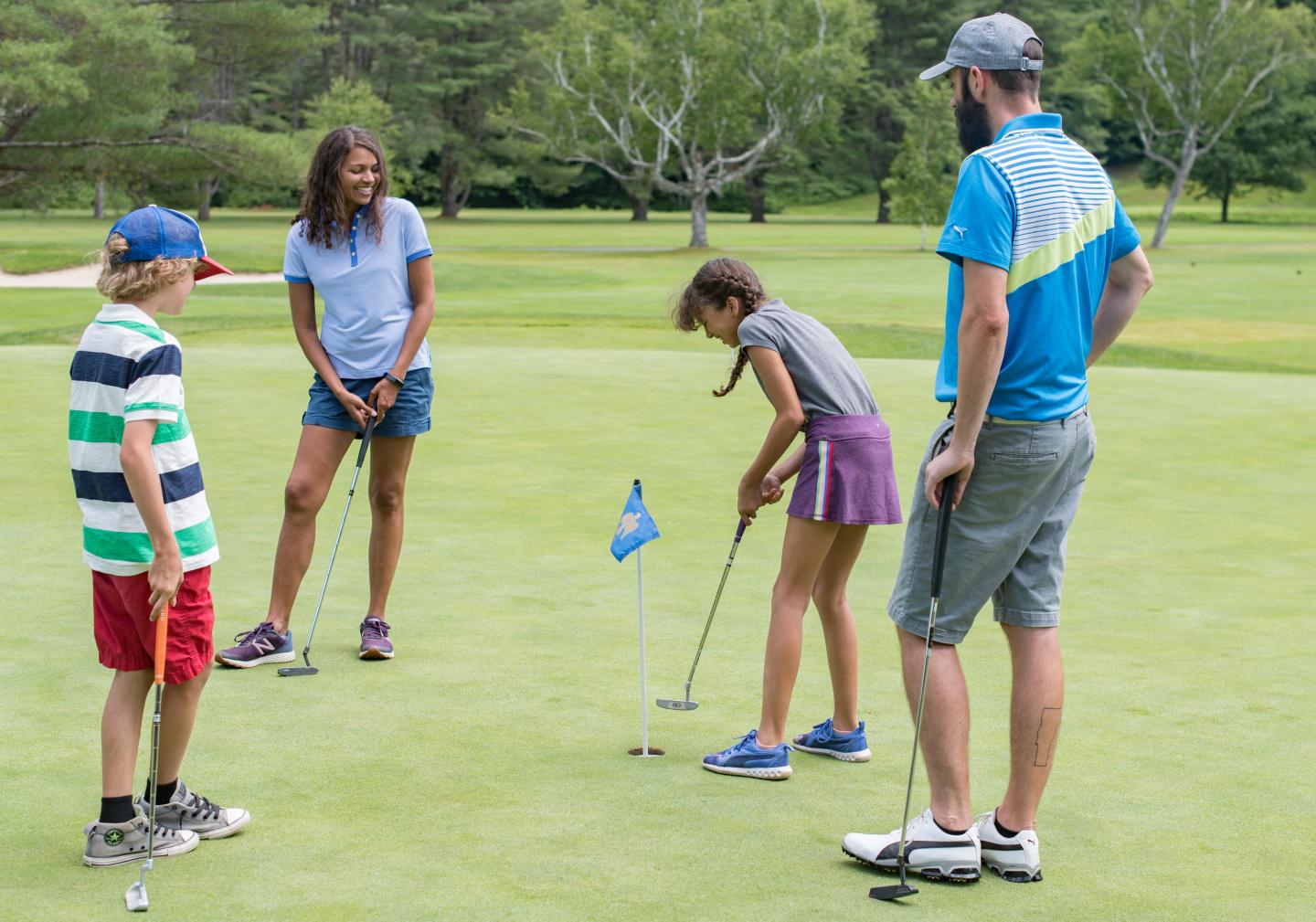 Girl playing mini-golf, watched by three people on green grass.