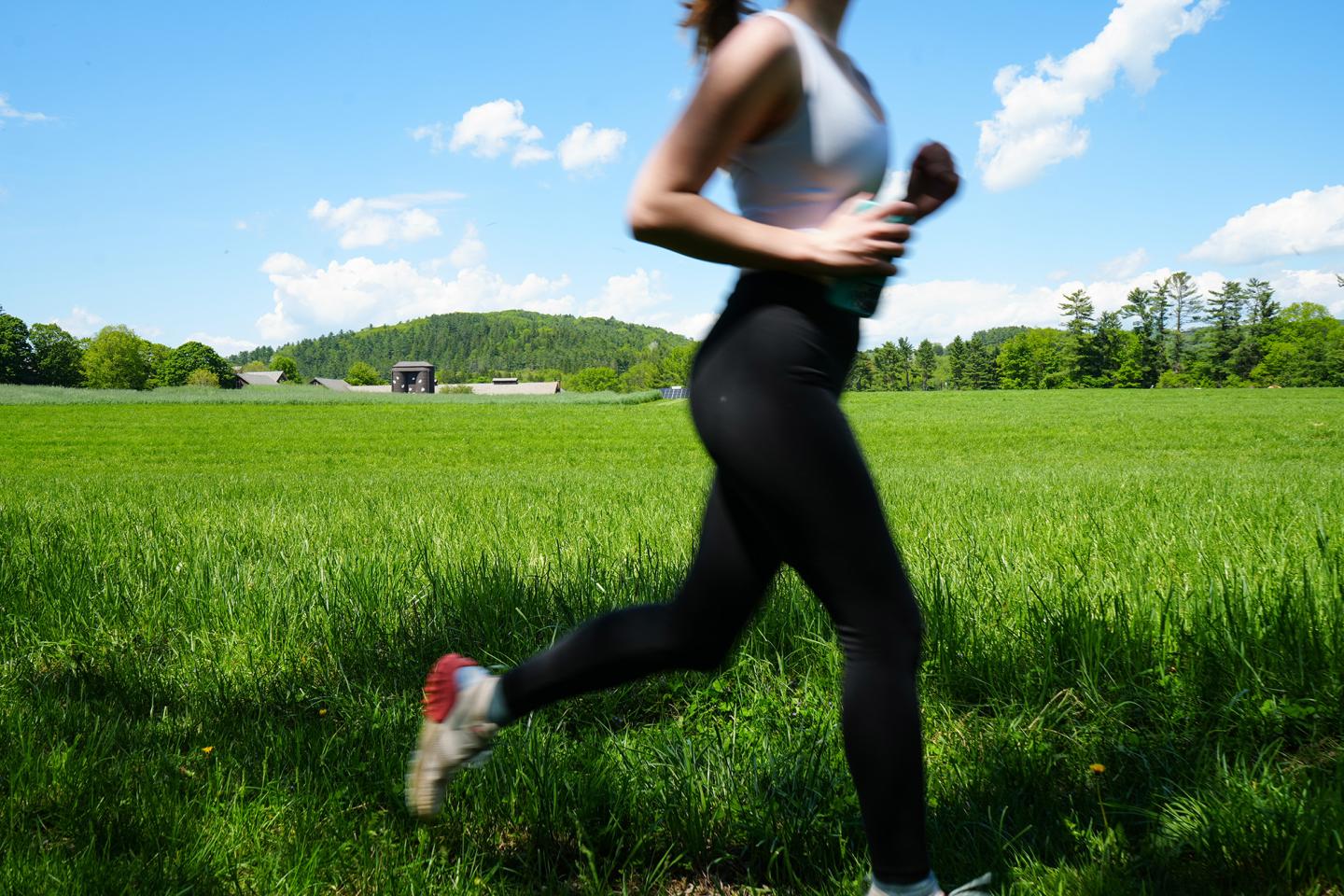 Woman jogging in a sunny, green field with a blue sky and clouds.