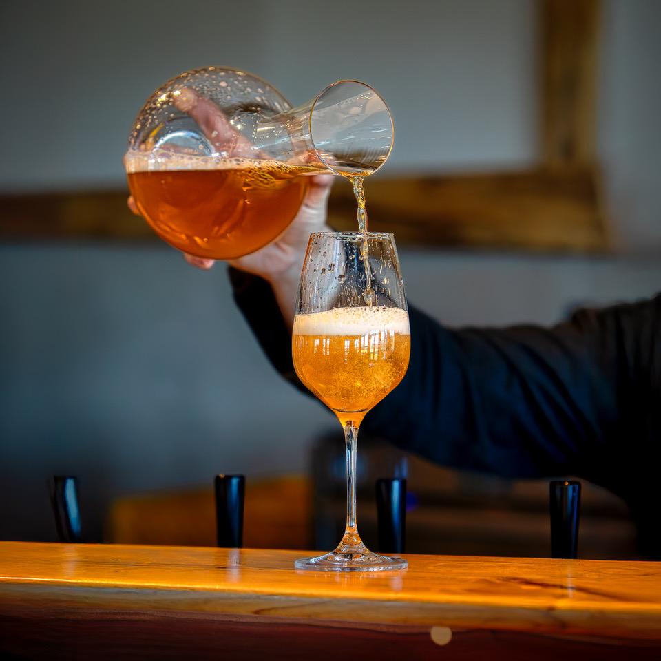 Pouring golden beer from a decanter into a glass on a wooden bar counter.
