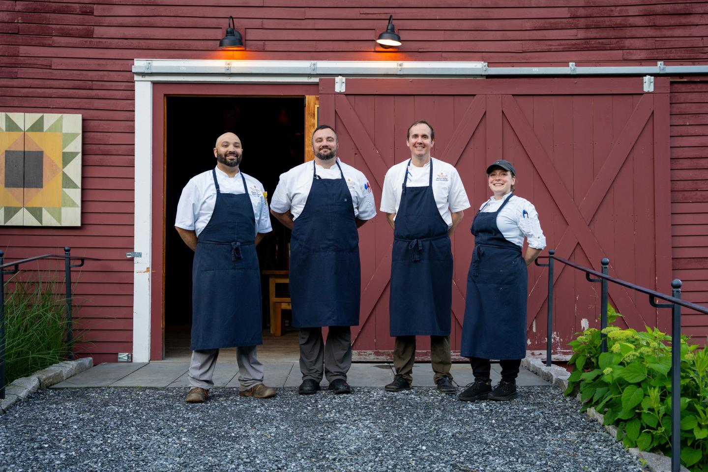 Chefs in aprons stand smiling in front of a red barn.