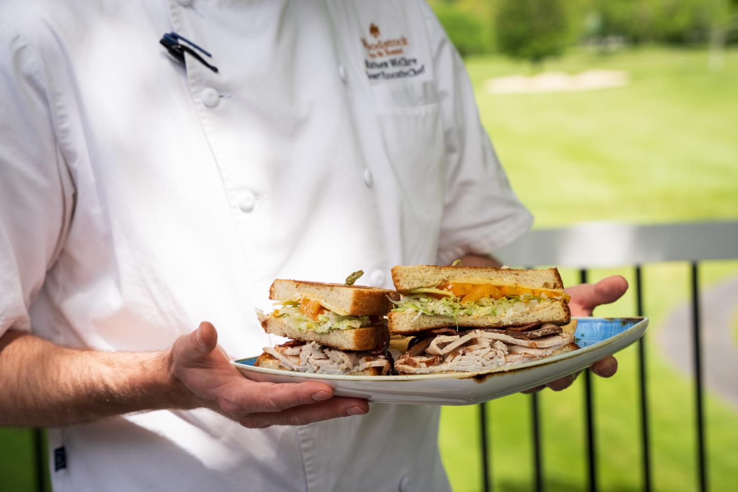 Chef holding a plate with stacked sandwiches in a sunny, outdoor setting.
