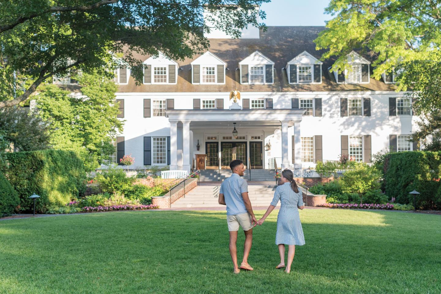 Couple holding hands, facing a large white building with green lawn and trees.
