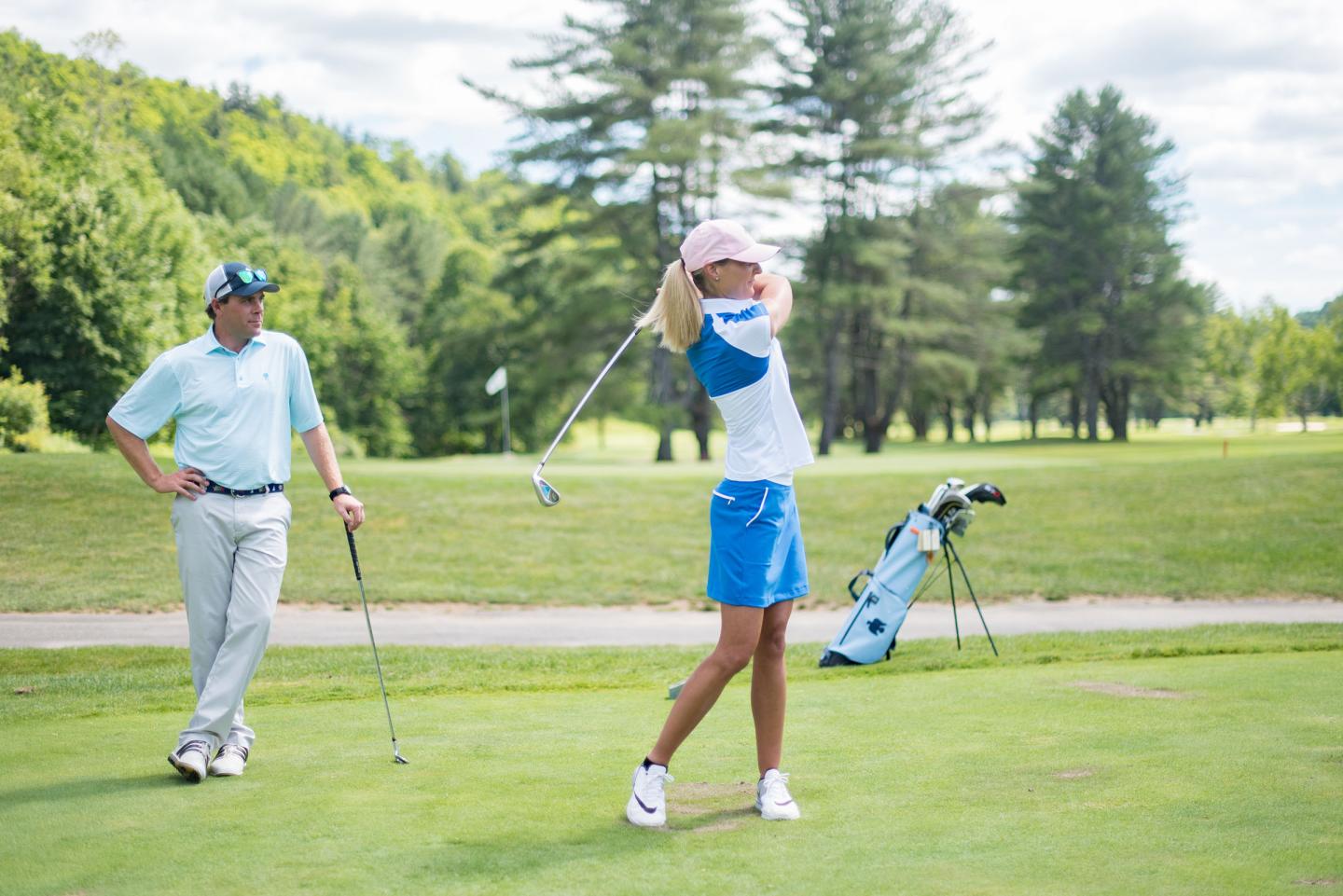 Golfer swinging club on lush green course; another person watches.