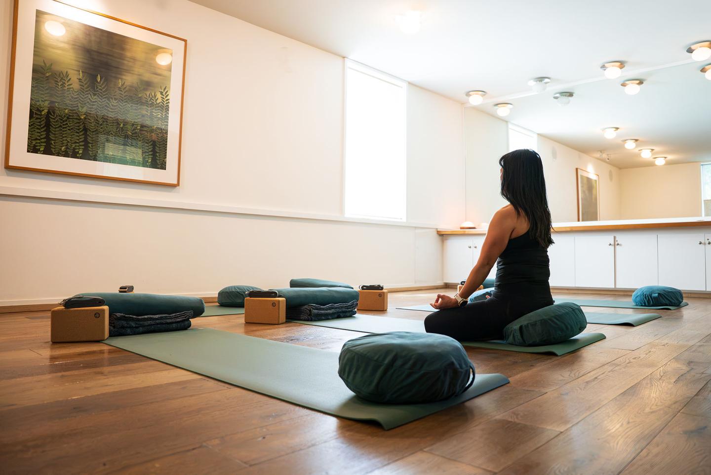 Woman meditating on yoga mat in bright studio with cushions and blocks.