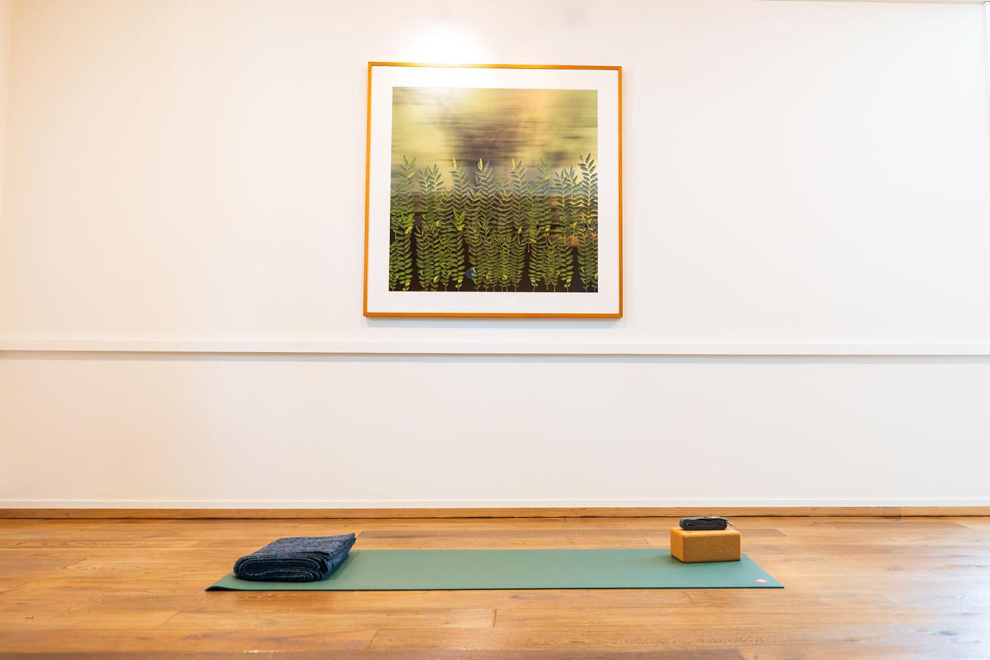 Yoga mat, blocks, and blanket on wooden floor; art on white wall.