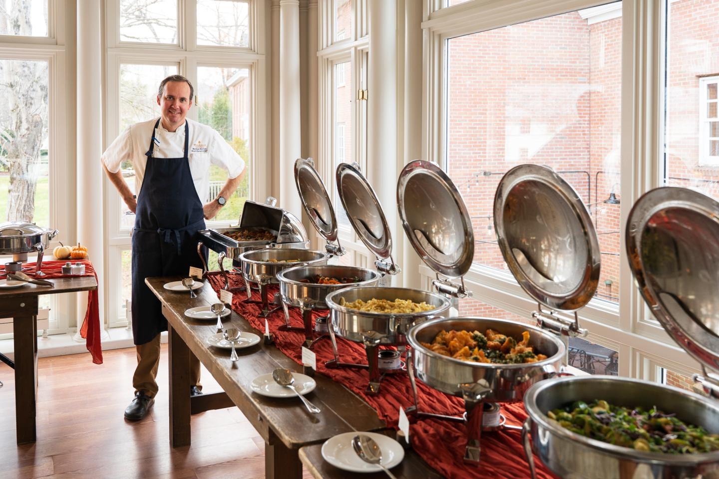 Chef stands by a buffet with various dishes in chafing dishes, near large windows.