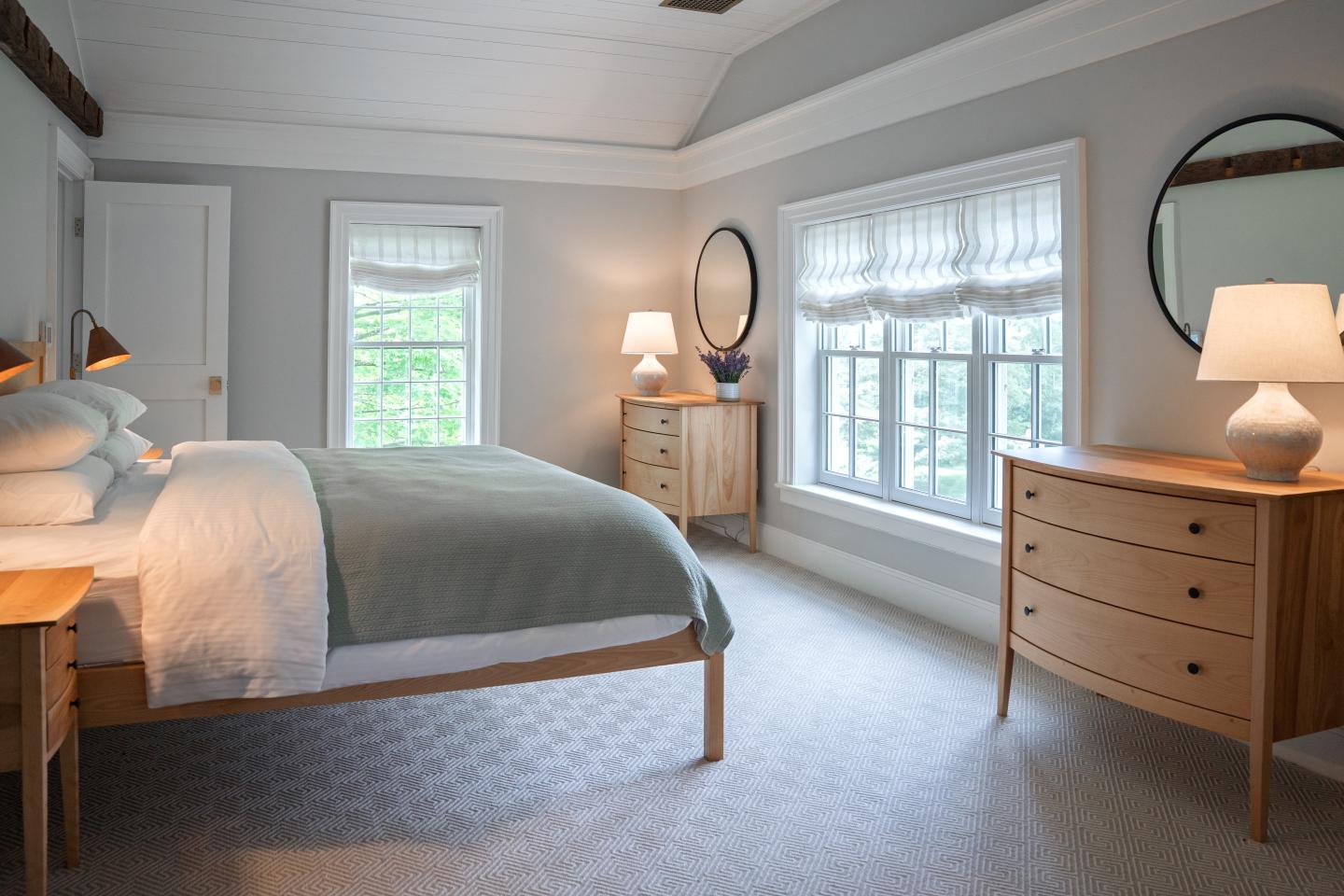 Bright modern bedroom with a wood bed, dresser, desk, and large windows.
