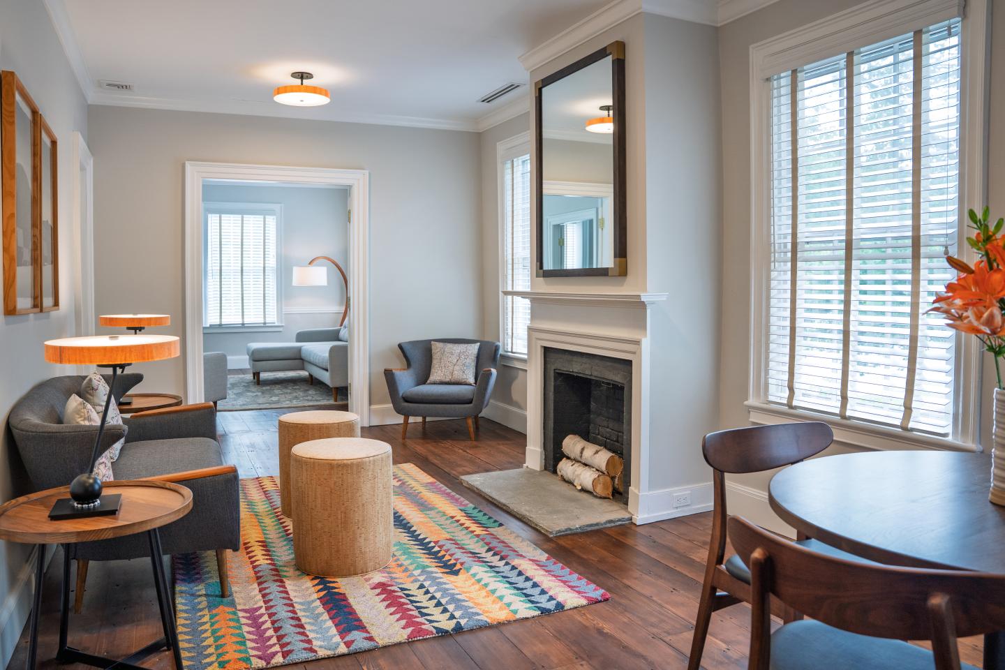 Bright, cozy living room with striped rug, fireplace, and large shuttered windows.