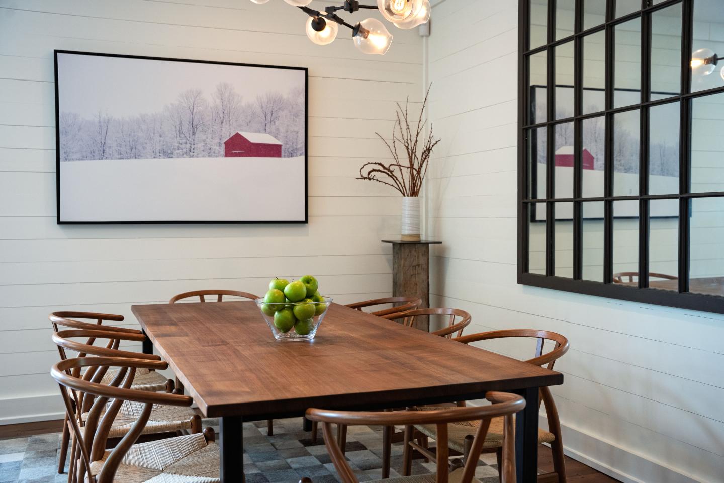 Modern dining room with wooden table, chairs, green apples, and large black-framed window.