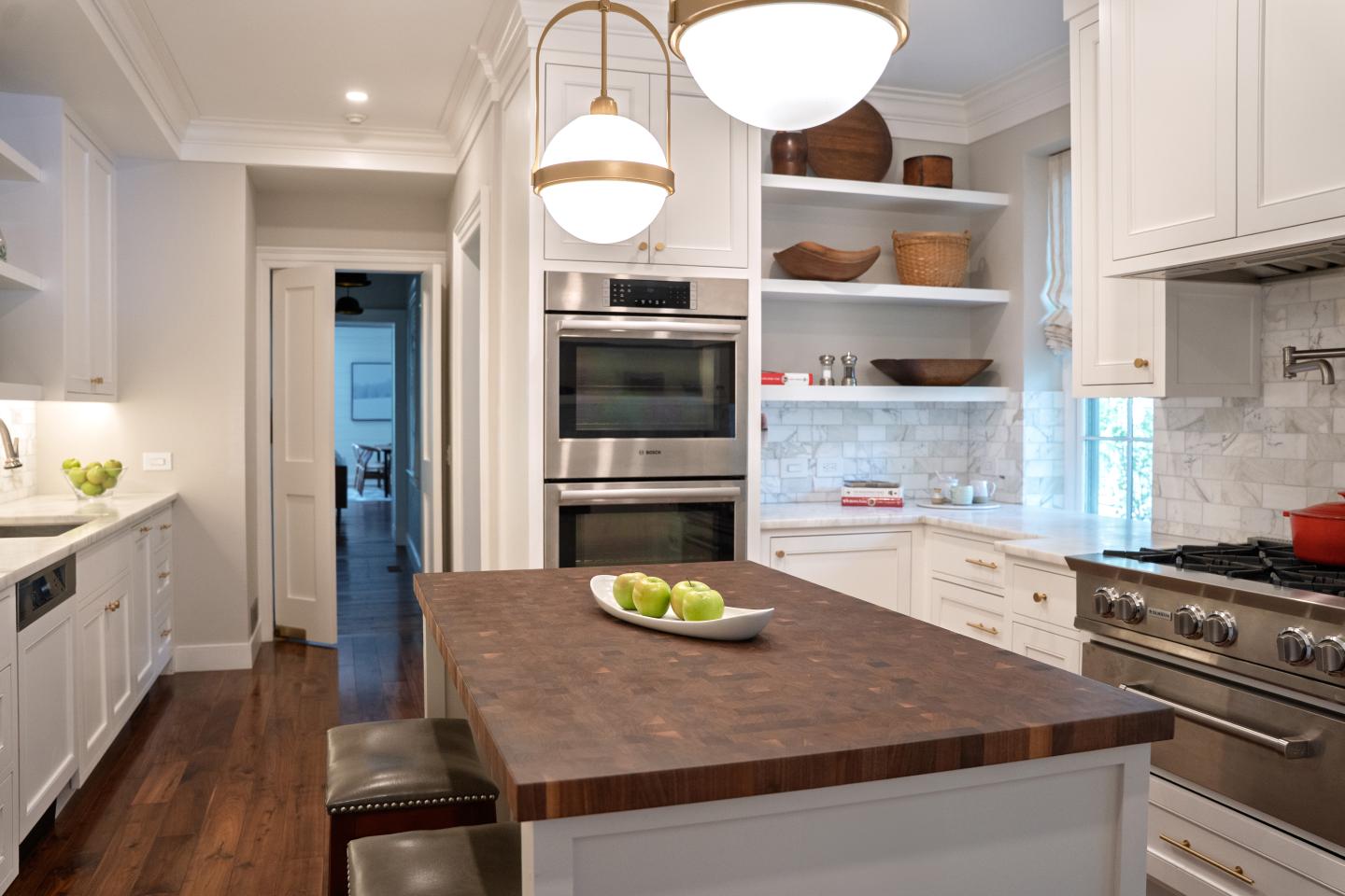 Bright white kitchen with a large wooden island, stools, and pendant lights.