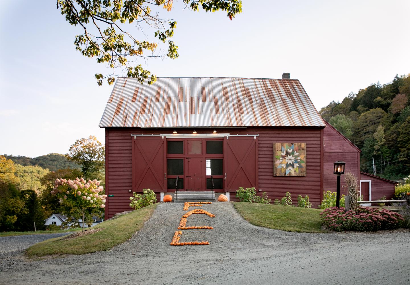 Red barn with rusted metal roof and autumn decorations beside a gravel path.
