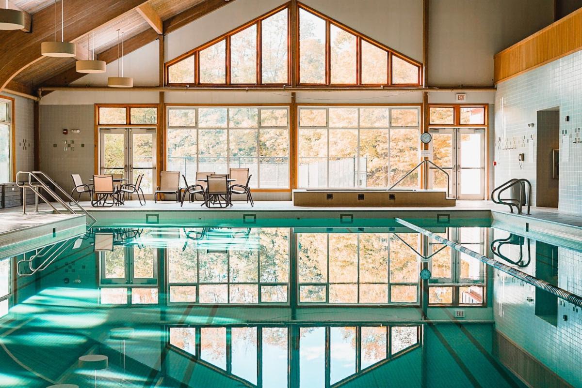 Indoor swimming pool with large windows and wooden beams reflected in turquoise water.