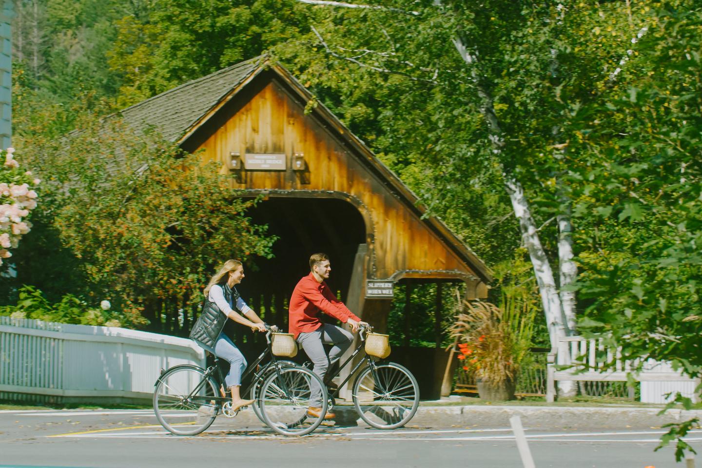 Two people cycling past a wooden cottage surrounded by green trees.