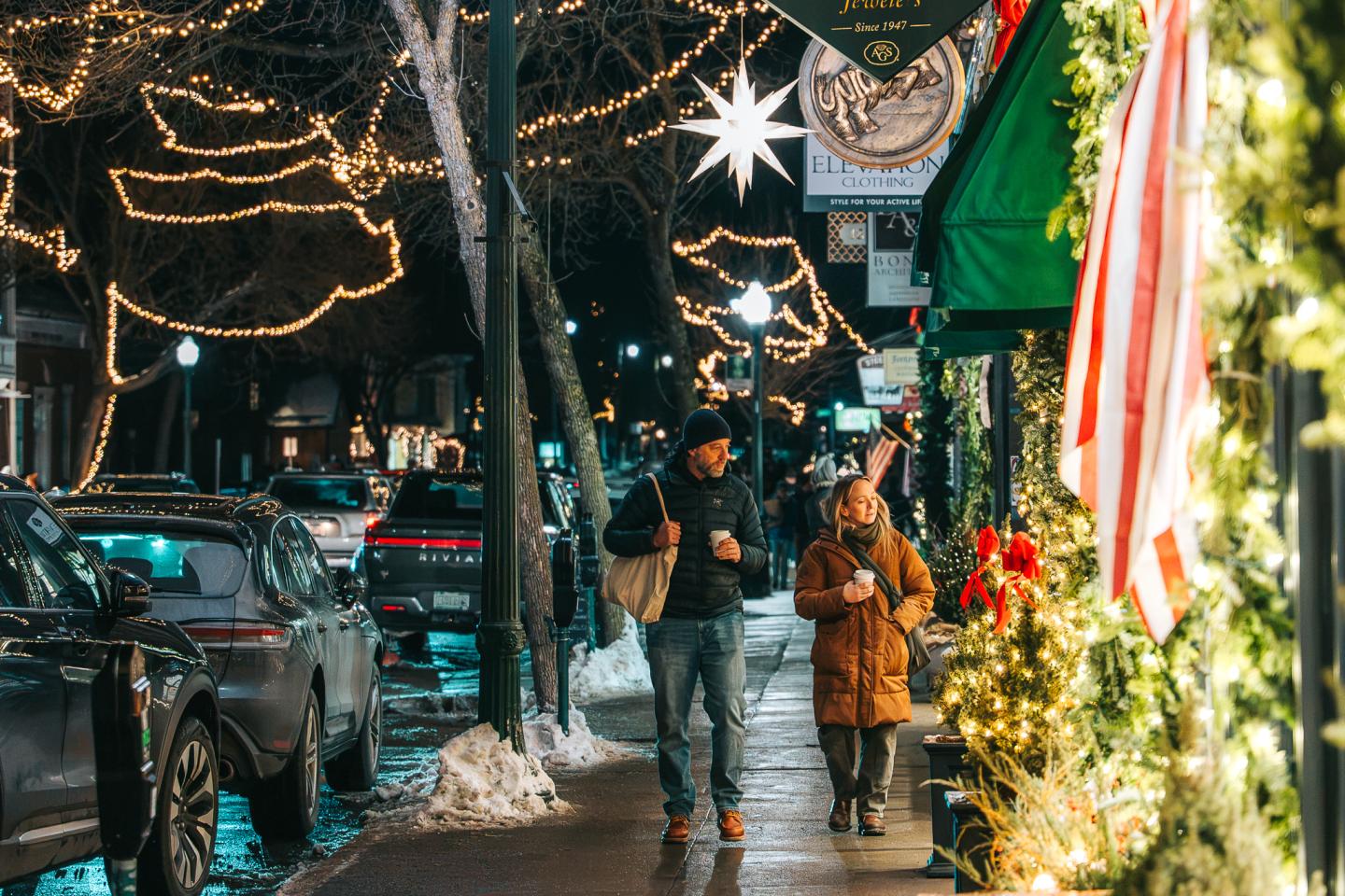 Holiday-decorated city street with pedestrians walking past shops and parked cars.
