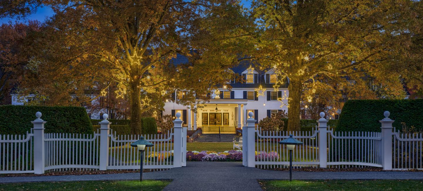 Grand white mansion framed by autumn trees and an iron gate at dusk.