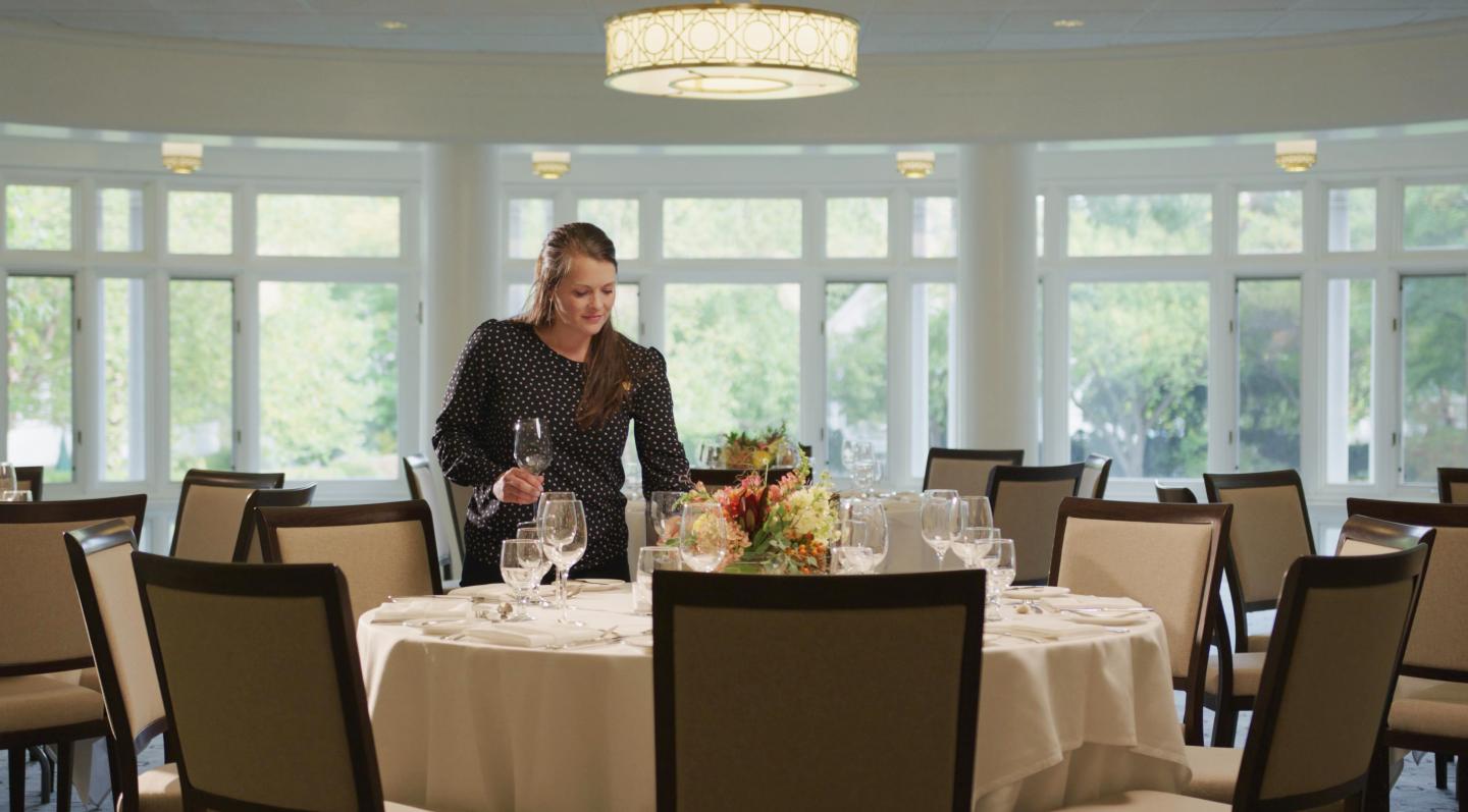 Woman arranging a floral centerpiece on a table in a bright dining room.